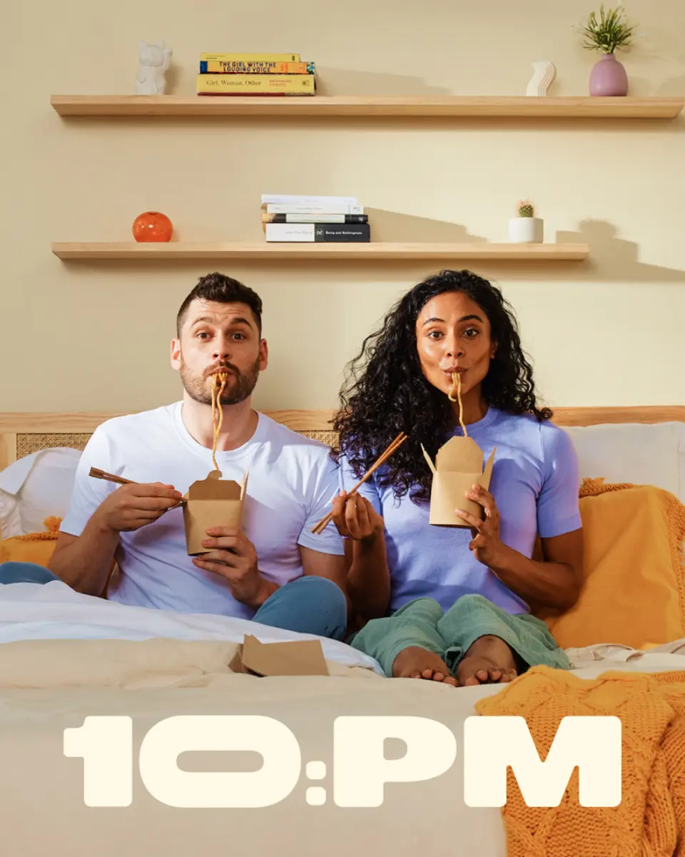 Couple sitting on a bed eating takeaway, with the 10:PM logo