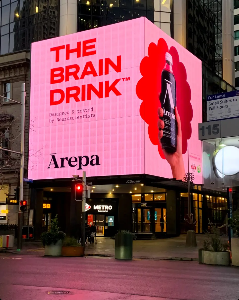 Ārepa "The Brain Drink" billboard on a building in a city square at dusk
