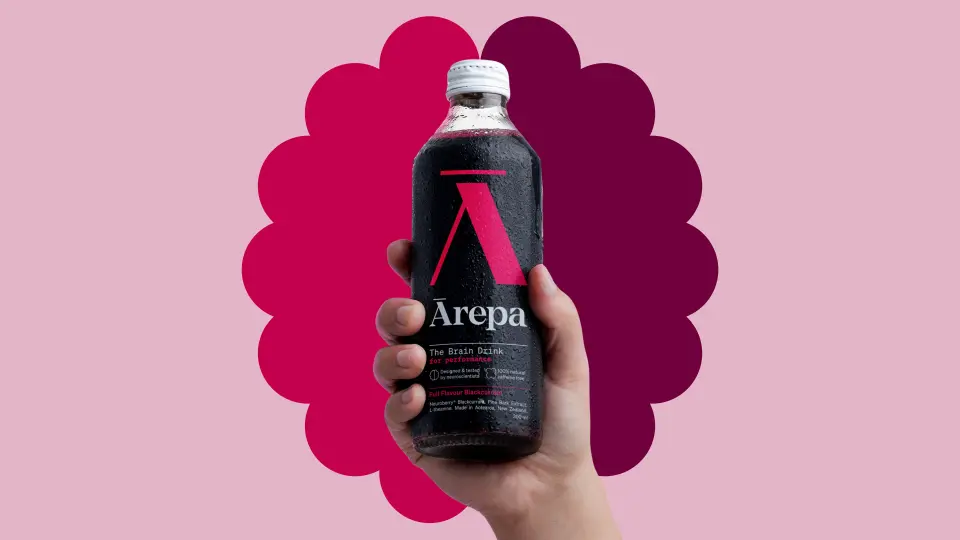 Hand holding an Ārepa bottle in front of a pink scalloped shape on a pale pink background