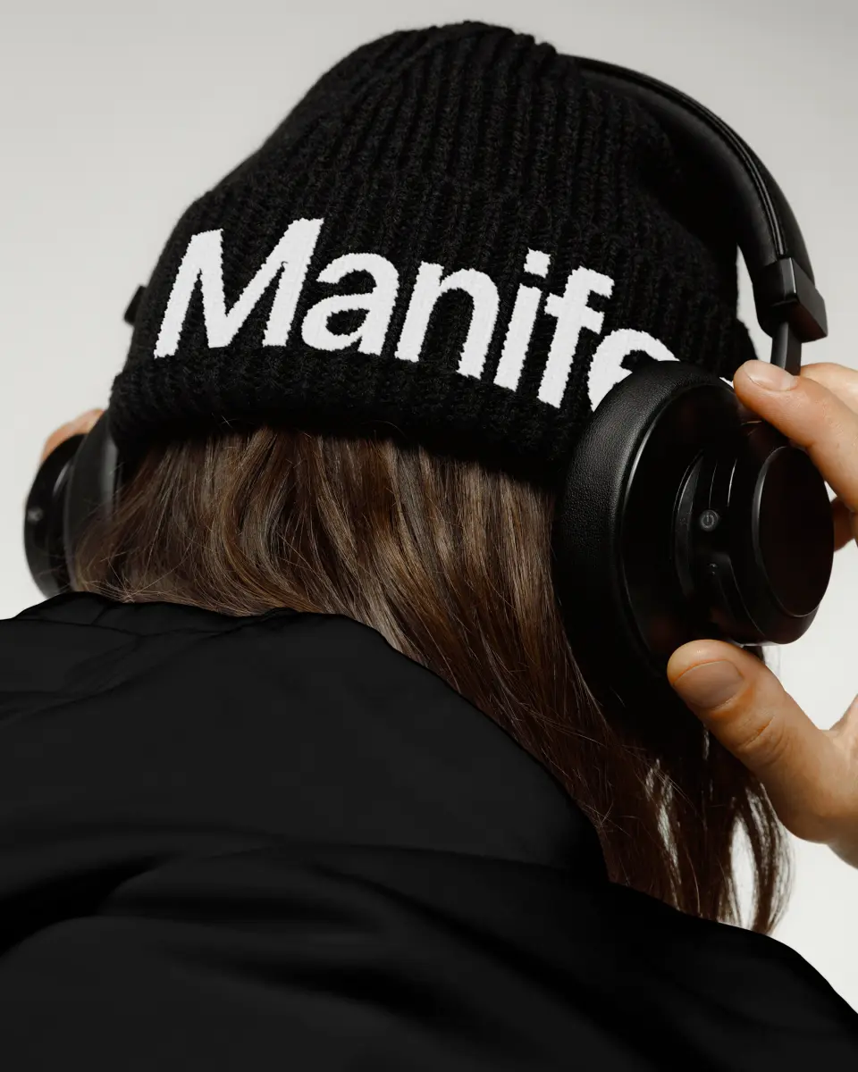 Person from behind wearing a black beanie reading Manifest and headphones