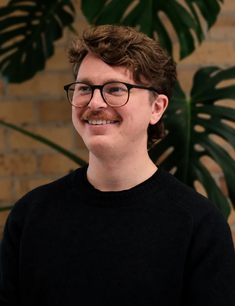 Portrait of Max Hurley, smiling in glasses and a black jumper in front of green plants